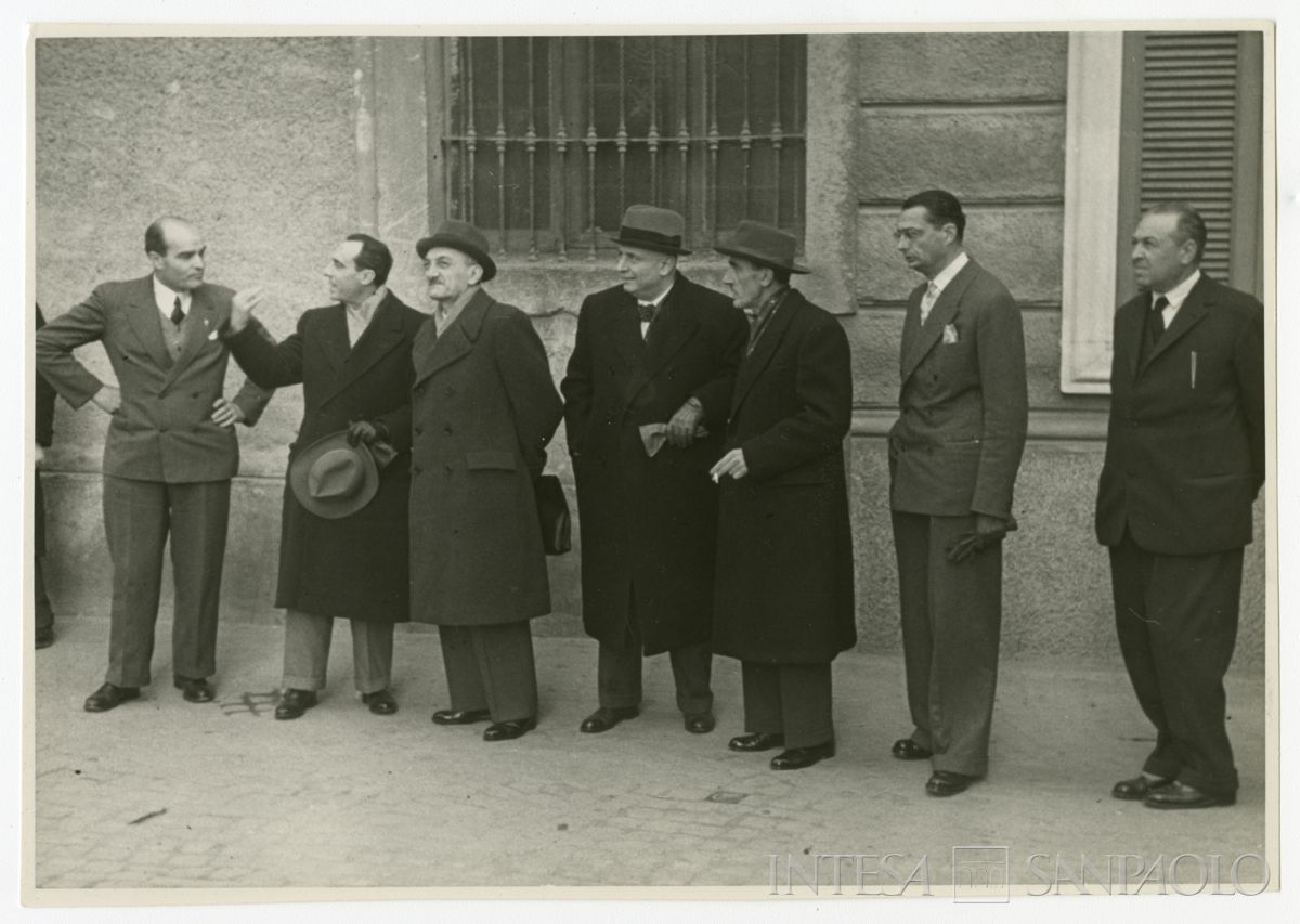 Autorità in riga lungo la parete dell'edificio prospiciente la filiale Cariplo di Lissone il giorno dell'inaugurazione; dal secondo da sinistra Bruno Minoja, Gian Luigi Dones, Stefano Jacini, persona n.i. e Sandro Candiani, gennaio 1950 (fotografo sconosciuto)