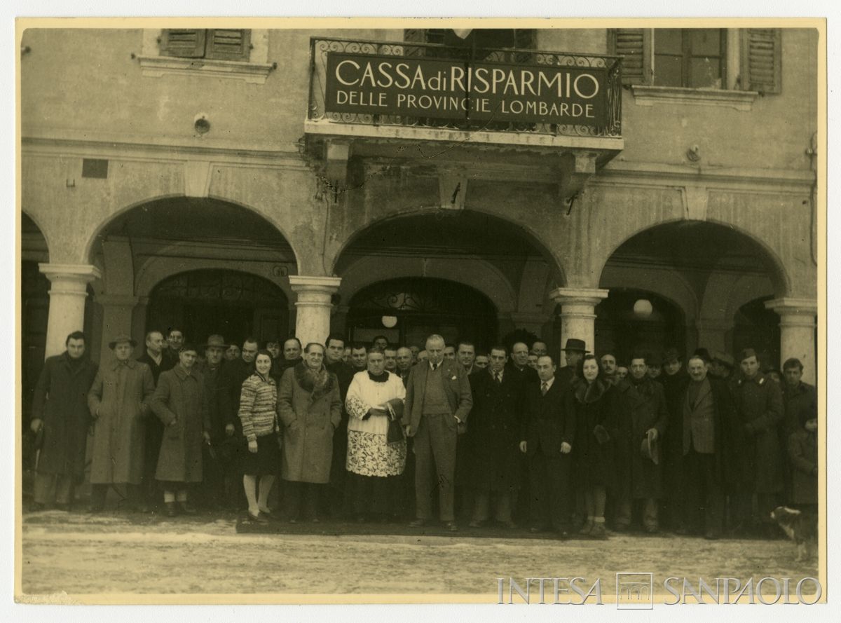 Foto di gruppo all'esterno della filiale Cariplo di Gonzaga il giorno dell'inaugurazione; in prima fila a destra del parroco il presidente Stefano Jacini, 10 gennaio 1947 (fotografo sconosciuto)