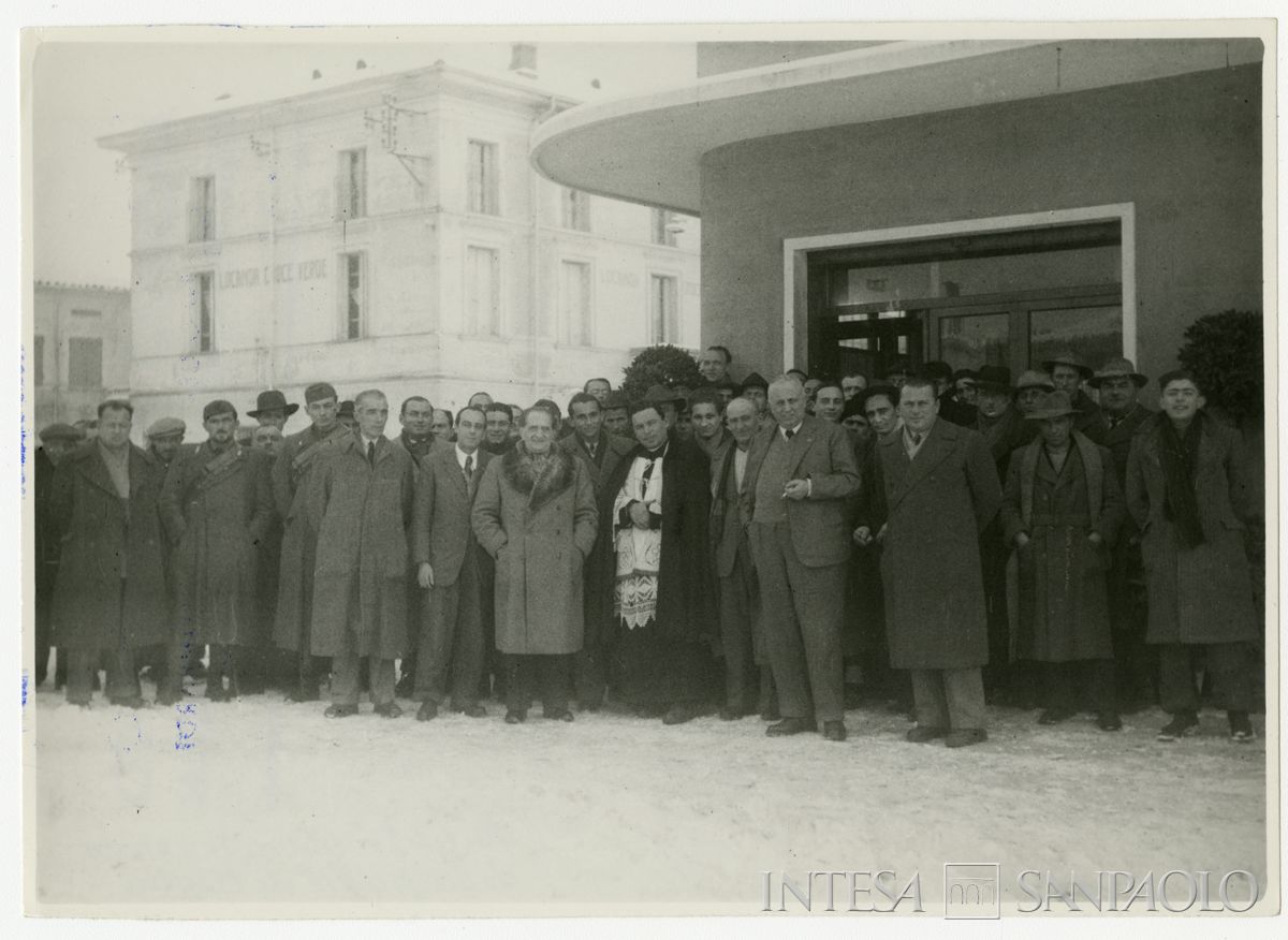 Foto di gruppo all'esterno della filiale Cariplo di Moglia il giorno dell'inaugurazione; terzo a sinistra del parroco Bruno Minoja, mentre a destra del parroco, davanti a tutti, con sigaretta in mano, è il presidente Stefano Jacini, 10 gennaio 1947 (fotografo sconosciuto)
