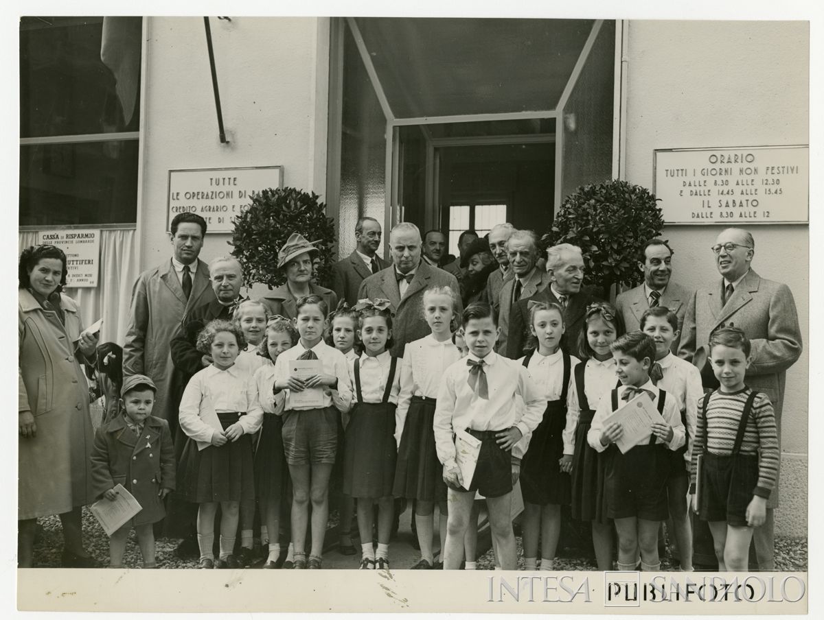Foto di gruppo di autorità e bambini all'inaugurazione dell'agenzia 22 di Milano della Cariplo, in via Jacopino da Tradate; secondo da destra, Bruno Minoja, il direttore generale Gian Luigi Dones e sulla stessa linea, poco distante, il presidente Stefano Jacini, 15 aprile 1950 (Publifoto)