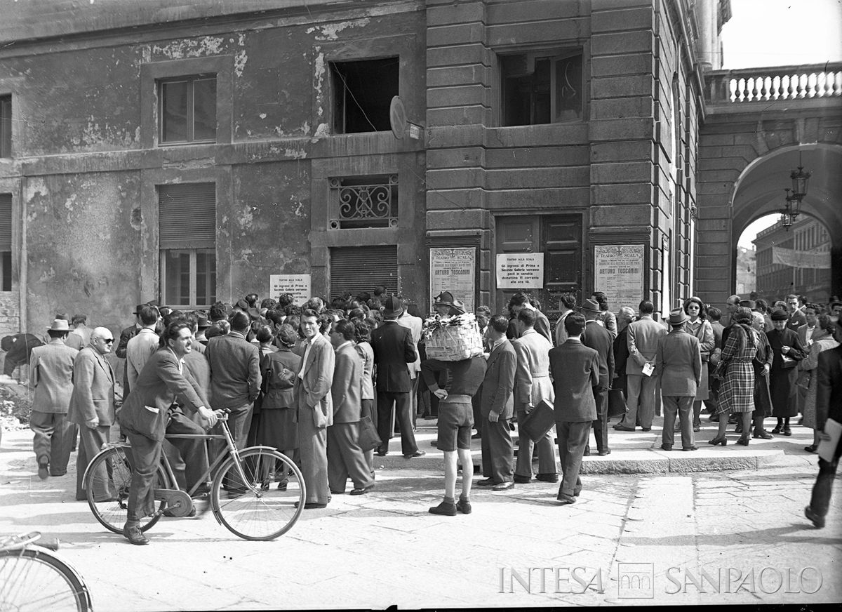 Un gruppo di persone staziona davanti ad una delle entrate laterali del Teatro alla Scala: visibili il manifesto del concerto di Arturo Toscanini per la riapertura e alcuni cartelli sulla vendita dei biglietti, 10 maggio 1946 (Publifoto)