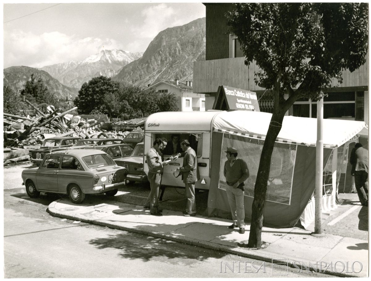 Lo sportello provvisorio della banca in una roulotte a Gemona, sullo sfondo le macerie di un edificio, post 6 maggio 1976. Archivio Storico Intesa Sanpaolo, sezione fotografica BCV