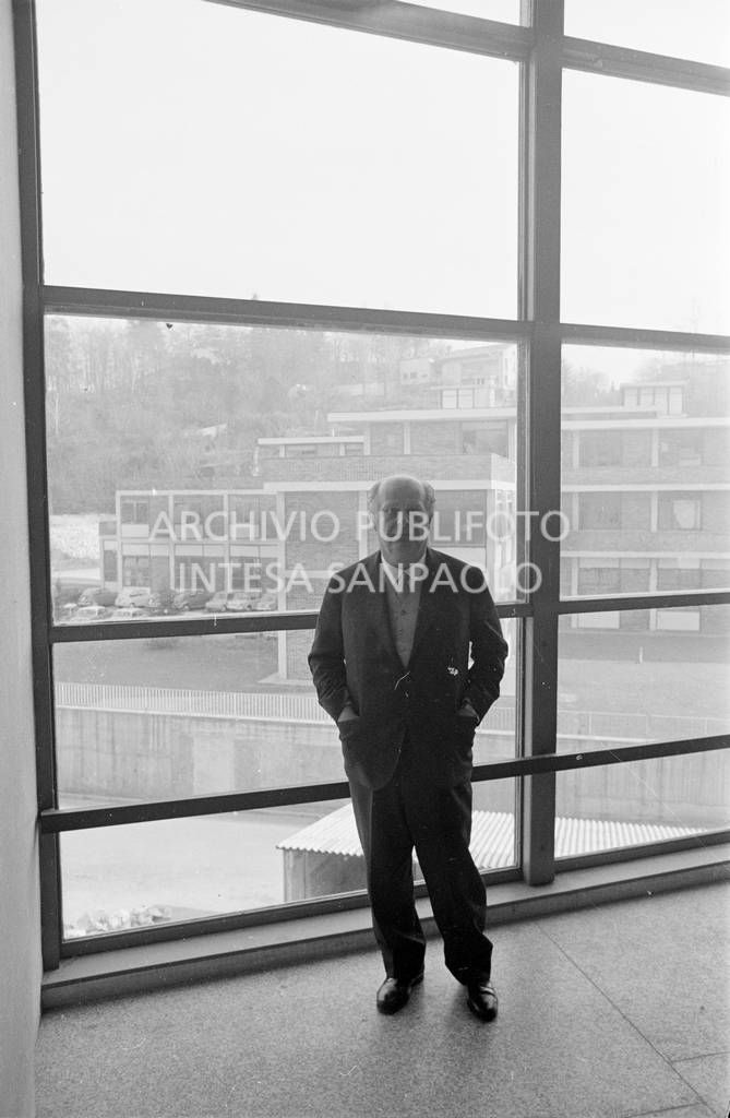 Adriano Olivetti ritratto di fronte a una vetrata; sullo sfondo gli edifici industriali<br>453516