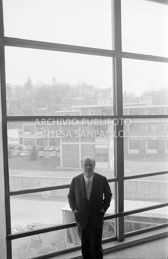 Adriano Olivetti ritratto di fronte a una vetrata; sullo sfondo gli edifici industriali<br>453517