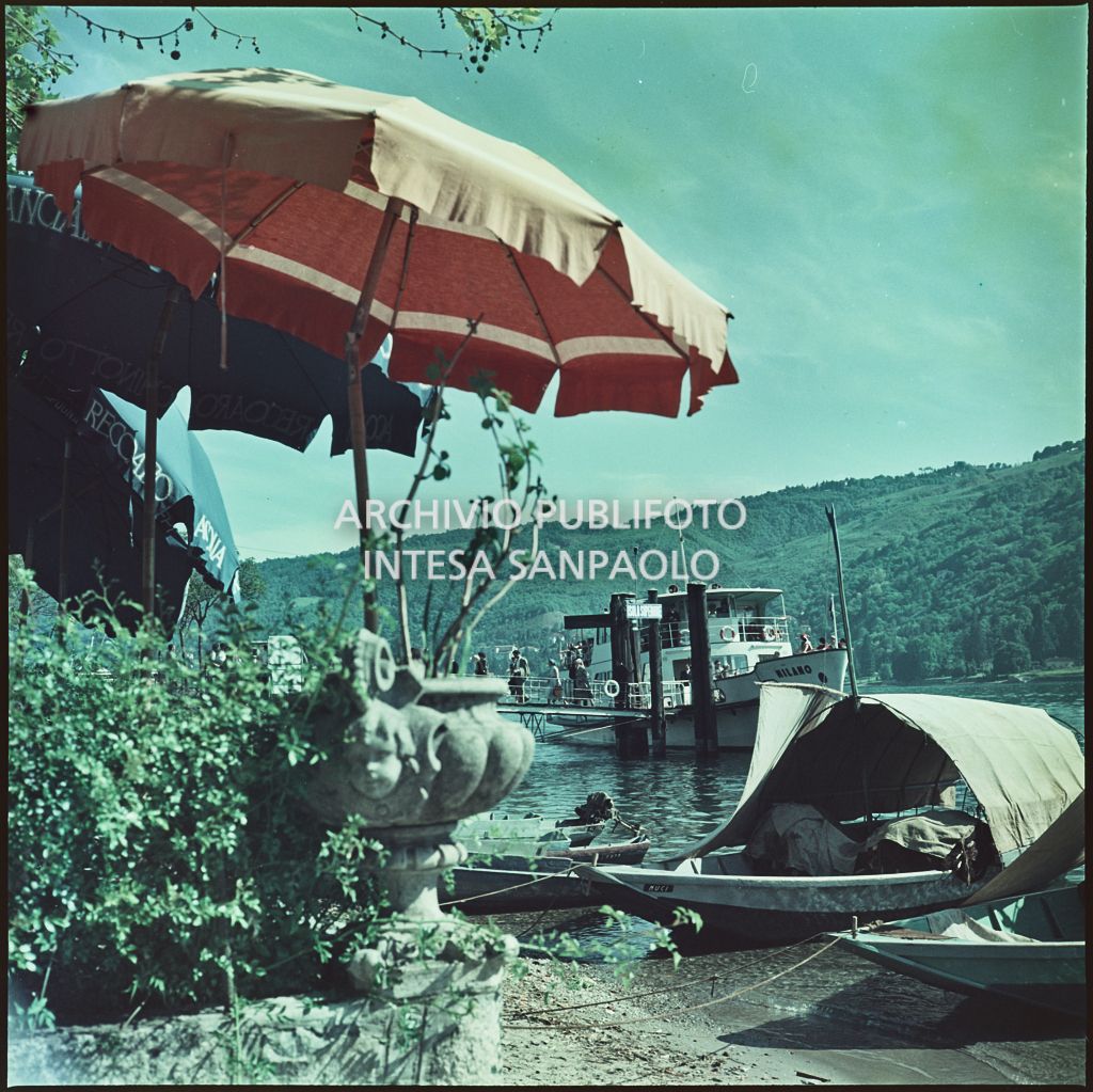 Vista dalla riva dell'Isola dei Pescatori sul lago Maggiore. In primo piano ombrelloni ed imbarcazioni, sullo sfondo un molo con attraccato un traghetto<br>335