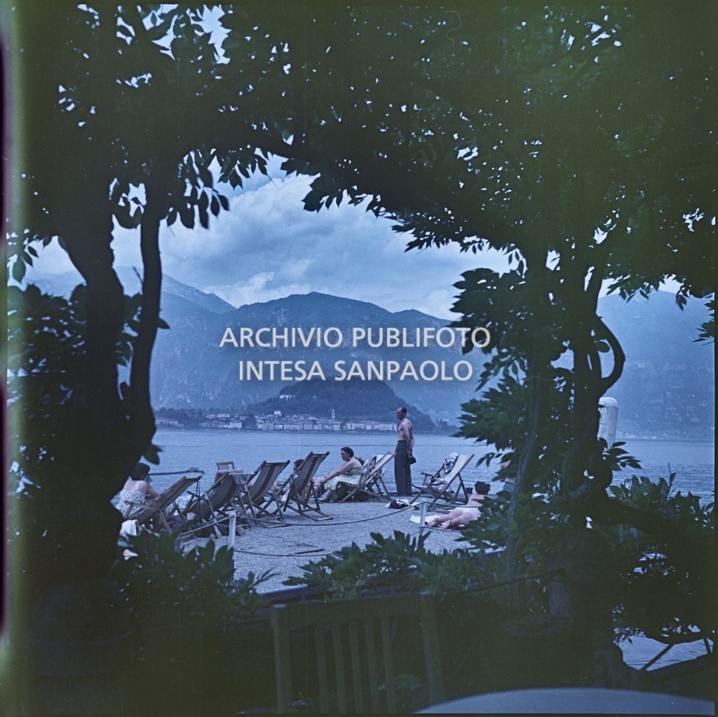 Turisti seduti sulle sdraio in riva ad un lago. In primo piano della vegetazione<br>337