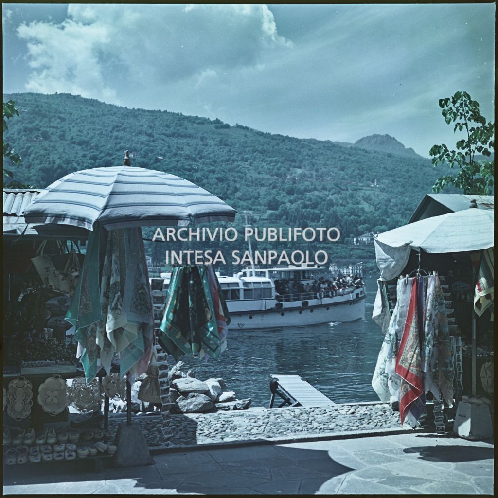 Bancarelle di souvenir sul lungolago. Sullo sfondo un traghetto e le montagne<br>344