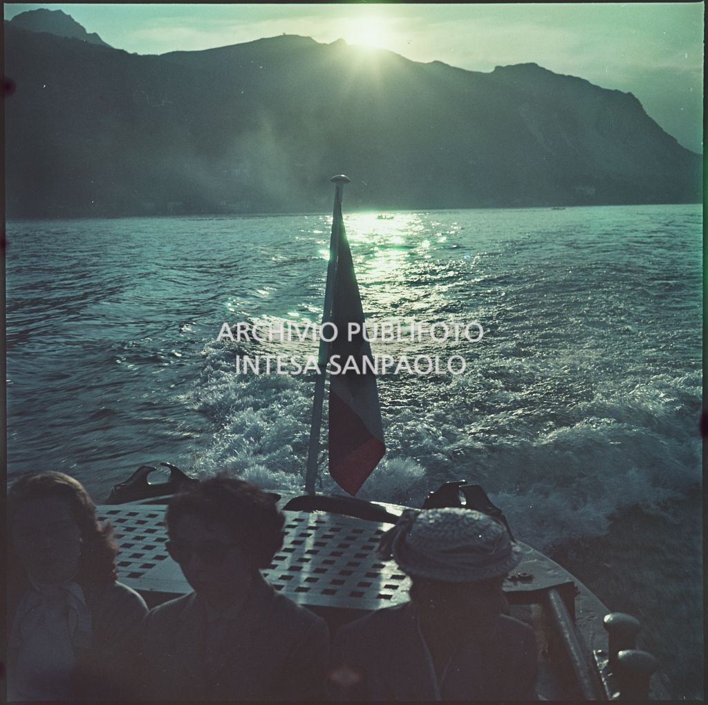 Vista del lago e delle montagne al tramonto. In primo piano tre donne sedute su imbarcazione con la bandiera italiana<br>345