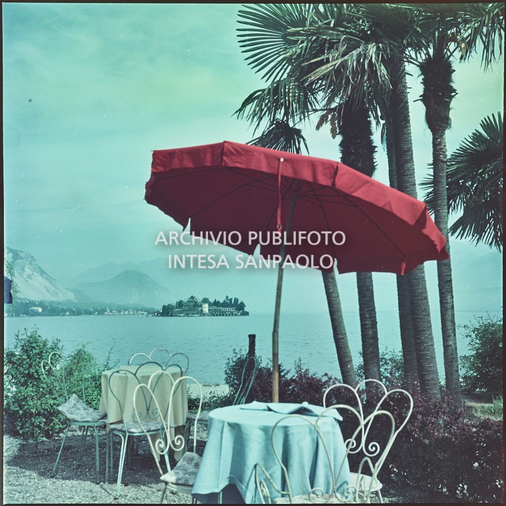 Tavolini ed ombrellone in riva al lago Maggiore. In lontananza è visibile l'Isola Bella<br>350