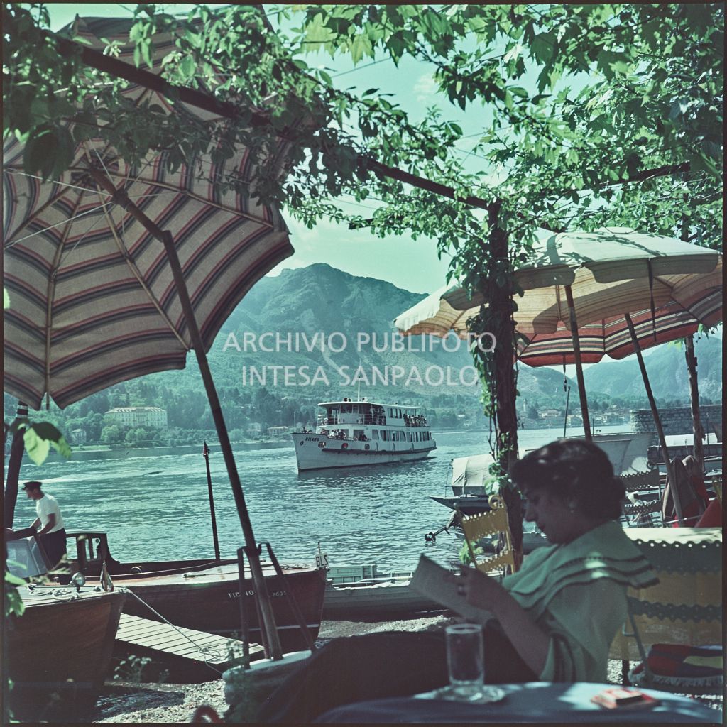 Vista del lago Maggiore e di un traghetto dalla riva dell'Isola dei Pescatori. In primo piano una donna legge un libro sotto ad un pergolato<br>352