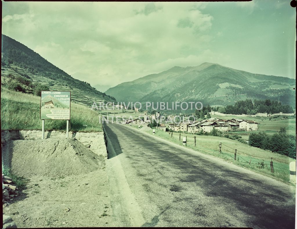 Vista su via Roma a Ponte di Legno. In lontananza è visibile il campanile della chiesa di San Giacomo, sullo sfondo le montagne, mentre sulla sinistra è presente un cartellone pubblicitario della scuola per bambini Kinderheim "I Folletti" di Ponte di Legno<br>1530