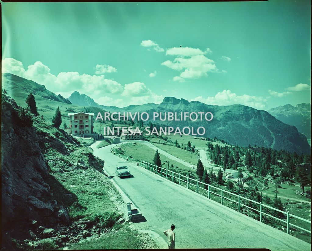 Strada di montagna sul Passo di Pordoi (Val di Fassa) con Hotel Pordoi e, più in basso, la relativa depandance e alberi. In primo piano un uomo sul ciglio della strada<br>2394