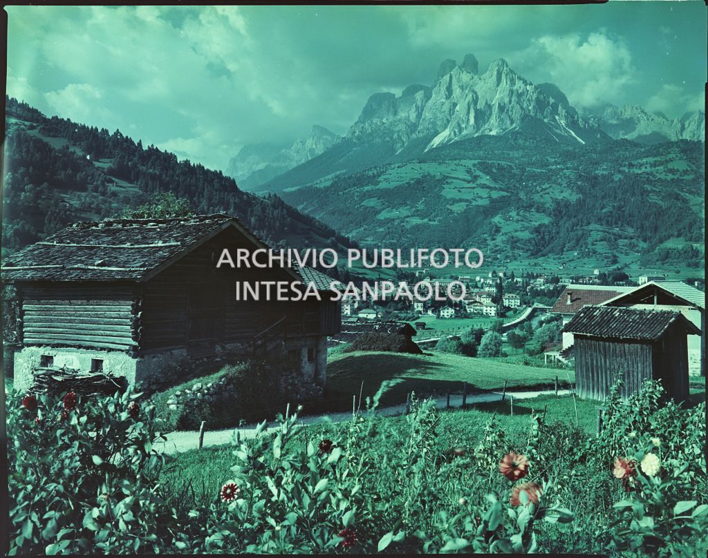 Vista su un paese in una valle. In primo piano una casa tipica in pietra e legno e sullo sfondo le Dolomiti<br>2395