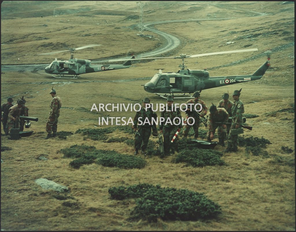 Gruppo di Alpini con tuta mimetica, cannoni e fucini. Sullo sfondo due elicotteri militari Agusta-Bell<br>5910