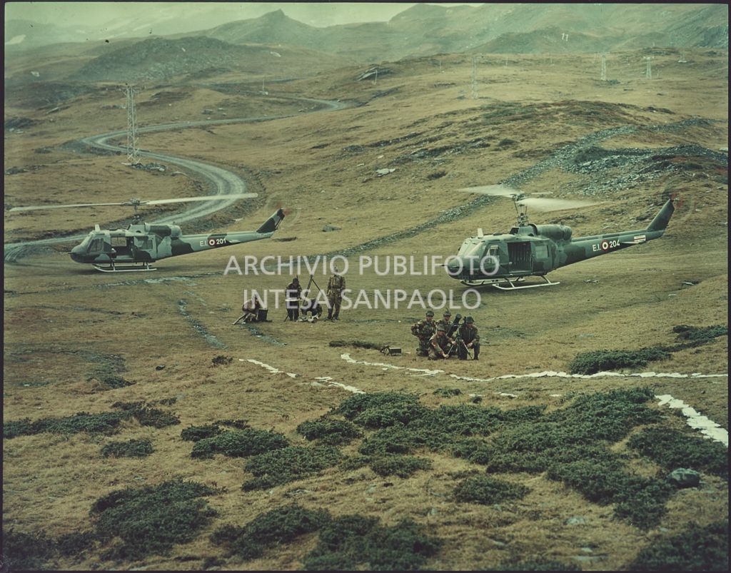 Gruppo di Alpini con tuta mimetica, cannoni e fucini tra le montagne. Sullo sfondo due elicotteri militari Agusta-Bell<br>5919