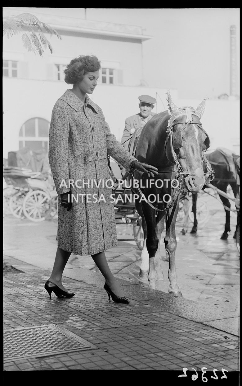 Indossatrice con cappotto si accosta ad un cavallo da traino con carrozza; sullo sfondo il conducente<br>22362