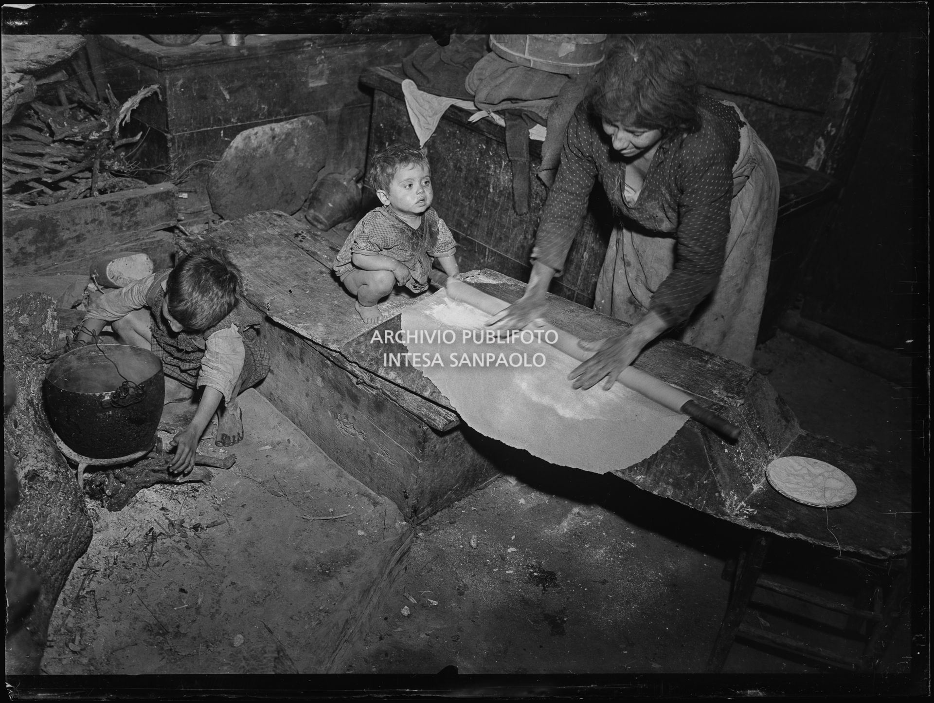 Africo, bambini e una donna che stende la pasta all'interno di una casa