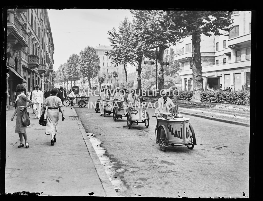 Venditori ambulanti di gelati  percorrono in colonna una strada di Milano a bordo delle loro biciclette con carretto