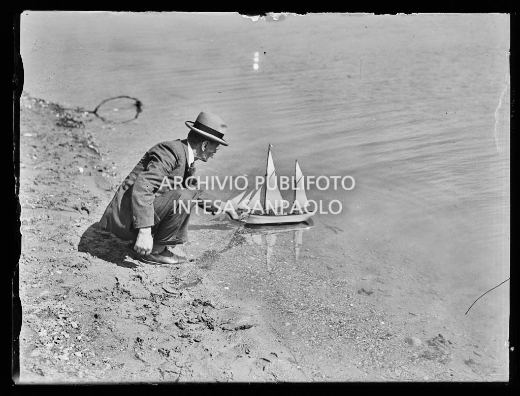Un uomo, in abiti eleganti, in riva al laghetto dei Giardini Pubblici di Milano, spinge in acqua il modellino di un veliero