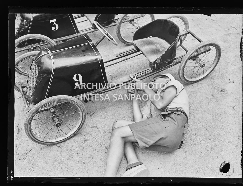 Un bambino, sdraiato a terra ai Giardini Pubblici di Milano, sistema con una chiave inglese un'automobilina a pedali