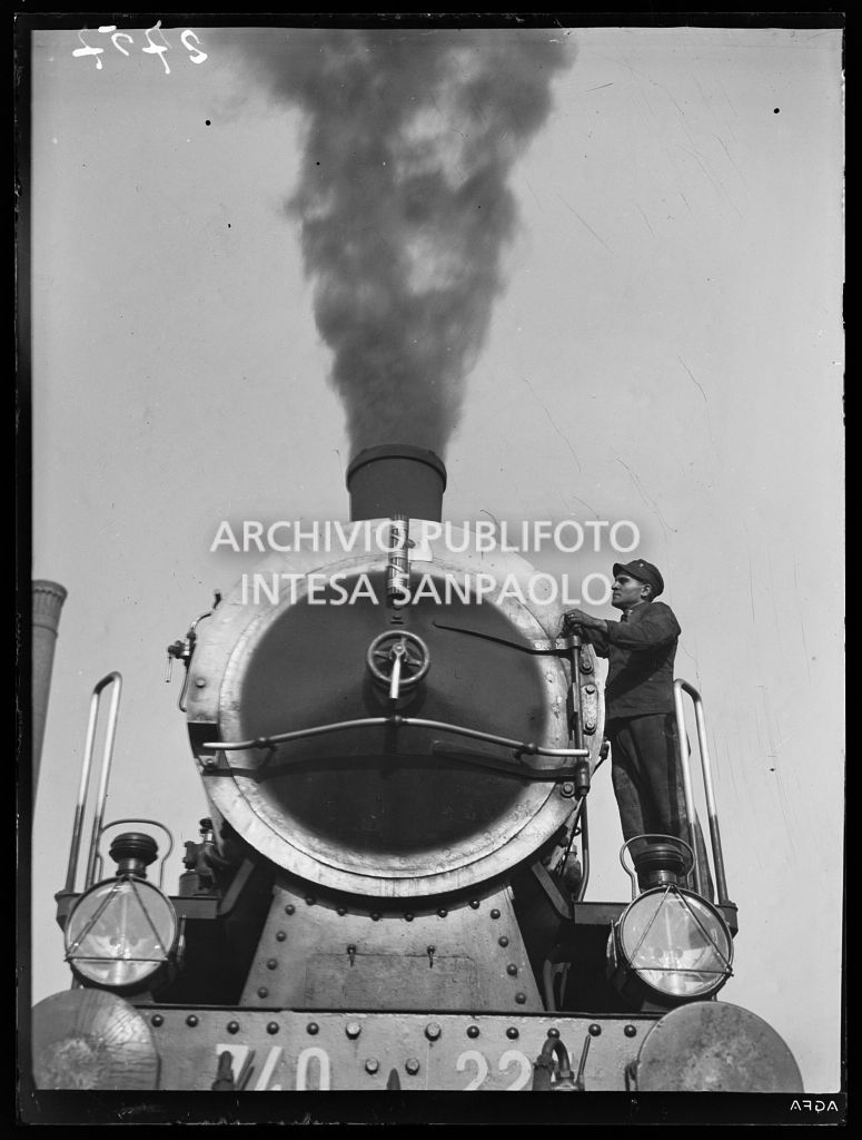 Macchinista in piedi su una locomotiva a vapore FS 740; dal camino uno sbuffo di fumo