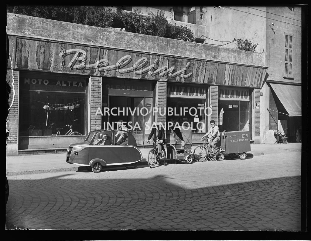 Tre modelli di bicicletta, adibite al trasporto di persone e merci, allineate con i loro conducenti davanti al negozio Bellini: su due di questi compare la sigla S.I.C.T. e il simbolo della città di Milano