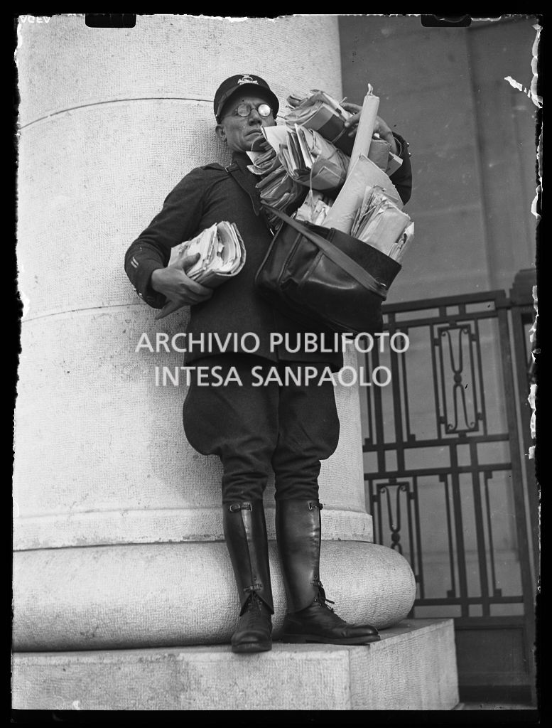 Un postino in divisa, con a tracolla la borsa straripante di corrispondenza, posa in piedi sulla base di una colonna
