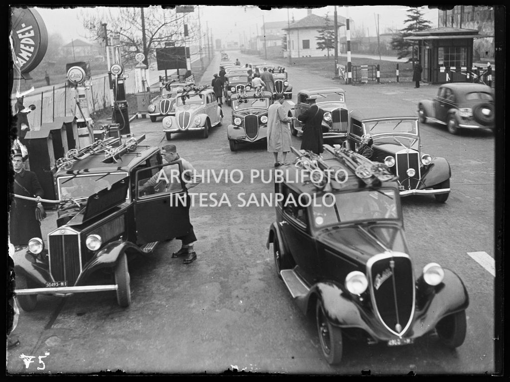 Automobili in coda, alcune di queste trasportano sul tetto degli sci; sulla sinistra una pompa di benzina Esso