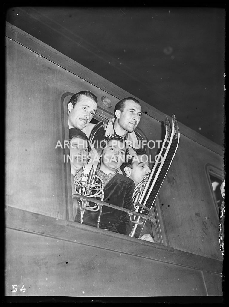 Un gruppo di sciatori posa affacciato al finestrino di un treno da cui spuntano gli sci