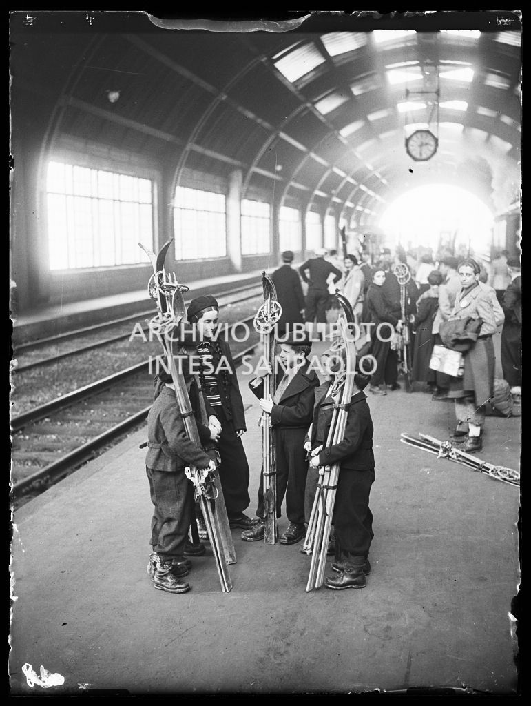 Bambini e ragazzi chiacchierano, imbracciando i loro sci, sulla banchina di una stazione, in attesa del treno