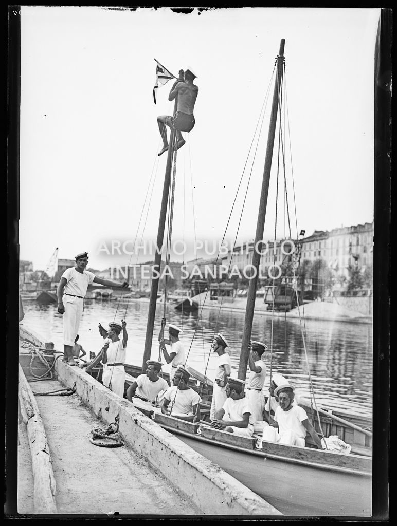 Giovani, in divisa marinara, a bordo di un bialbero con vela latina, attraccato alla Darsena di Porta Ticinese a Milano; uno di loro, arrampicato su un albero, issa una bandiera