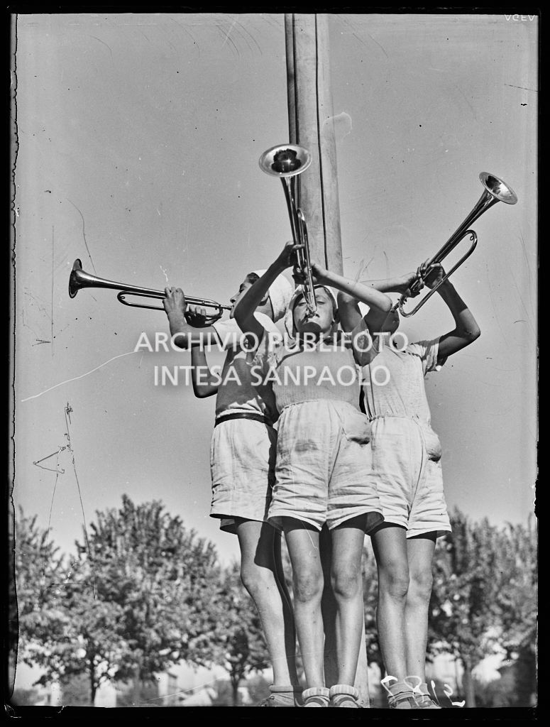 Tre bambini suonano la tromba ai piedi del palo alzabandiera in una colonia elioterapica