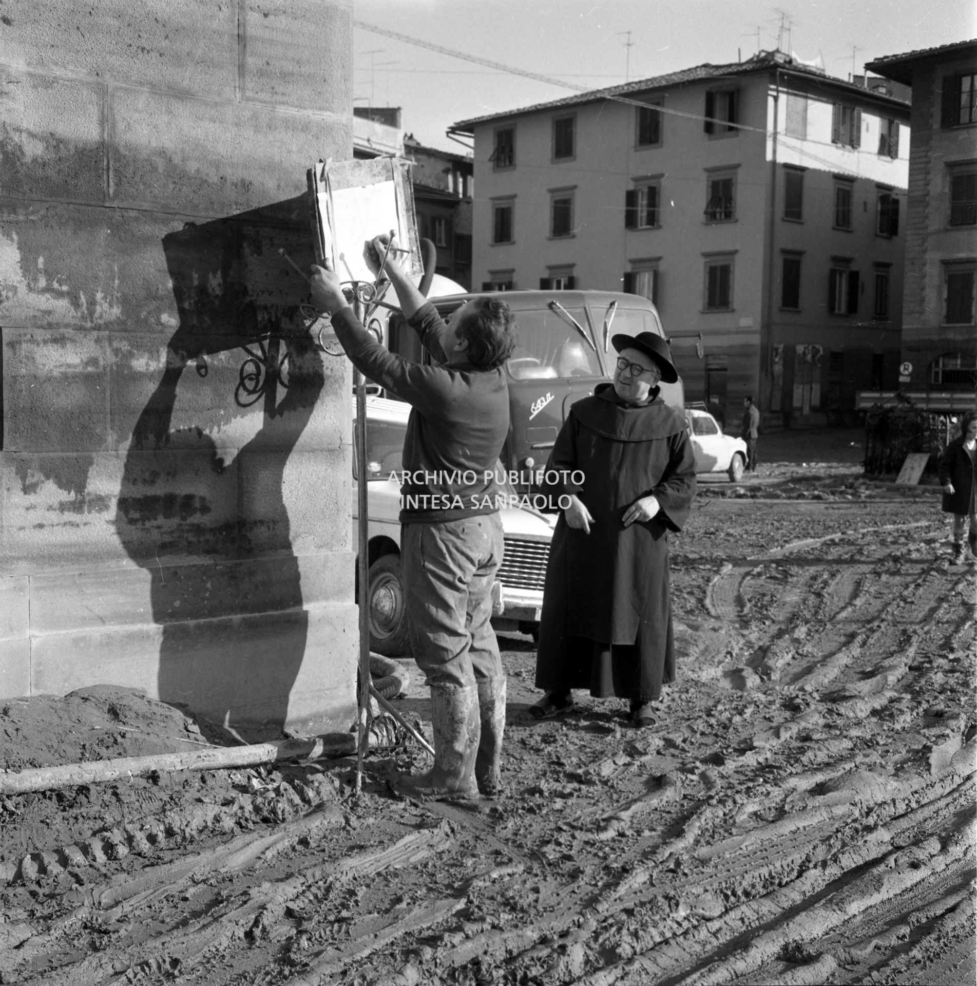 Un uomo affigge un cartello, osservato da un prete, in una via di Firenze dopo lo straripamento del fiume Arno
