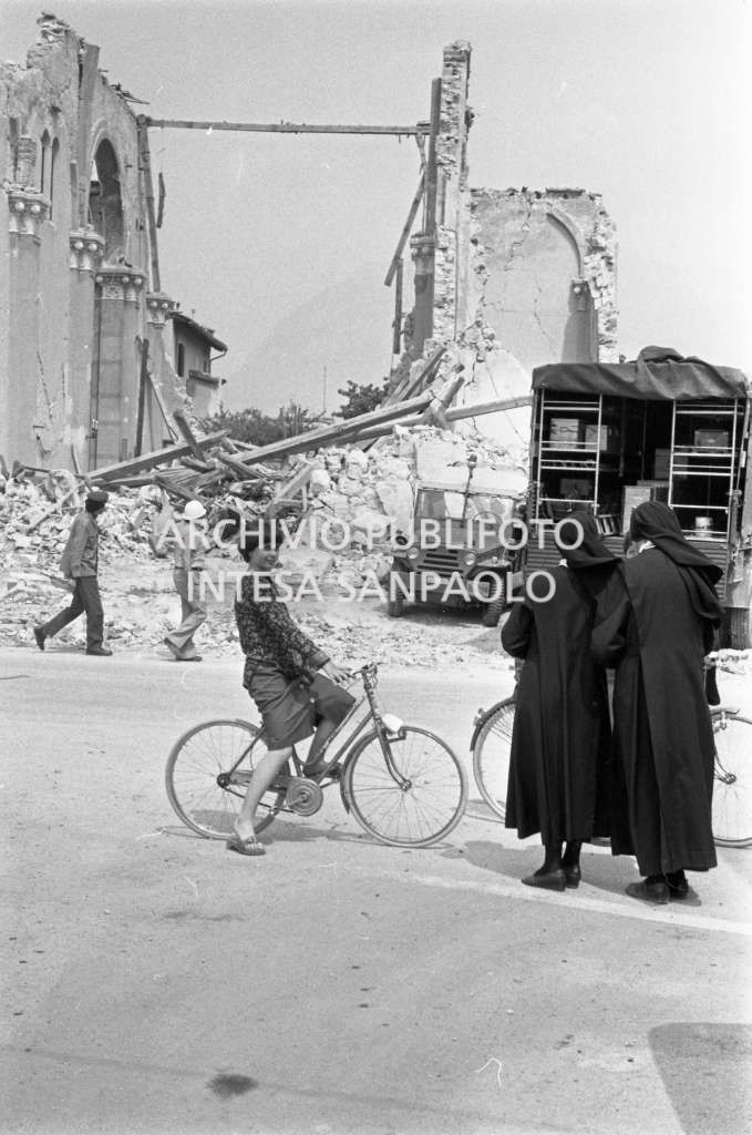 Due suore e una donna in bicicletta ritratte in una località colpita dal terremoto in Friuli, in secondo piano sono visibili i resti di una chiesa