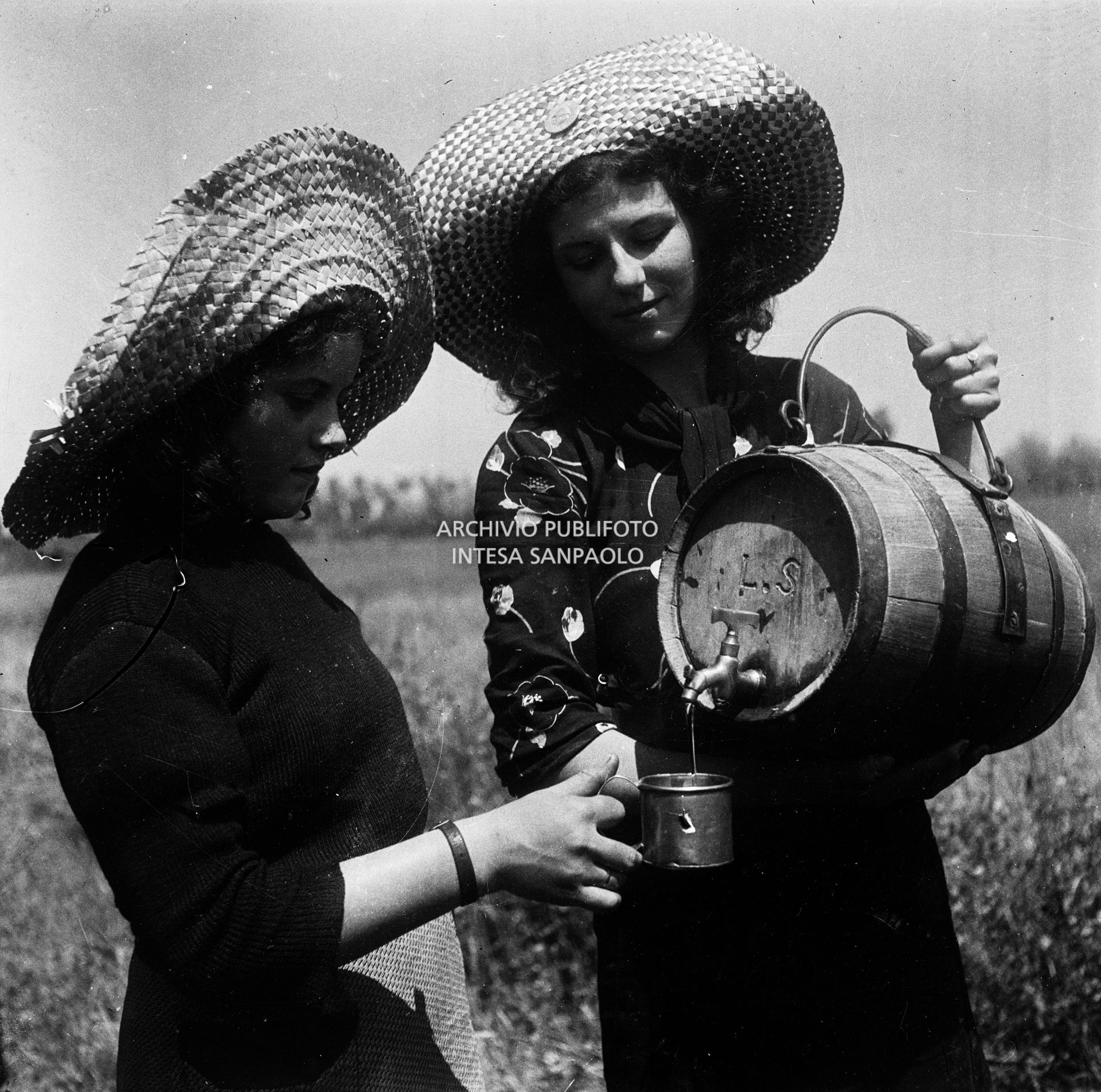 Due mondine ritratte in risaia mentre bevono da una piccola damigiana