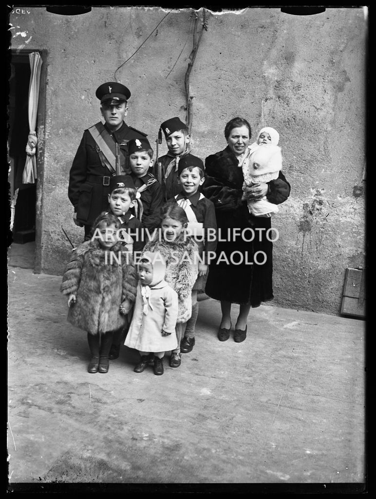 Una coppia, con i suoi otto figli, ritratta davanti all'entrata di casa; i figli maschi indossano divise da balilla e due di loro hanno il moschetto