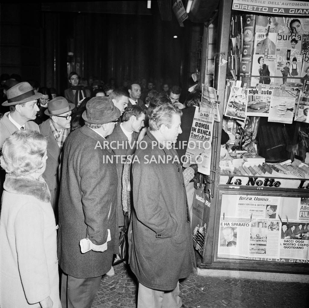 Folla davanti a un'edicola che espone i quotidiani che riportano la notizia dell'attentato alla Banca Nazionale dell'Agricoltura in piazza Fontana a Milano
