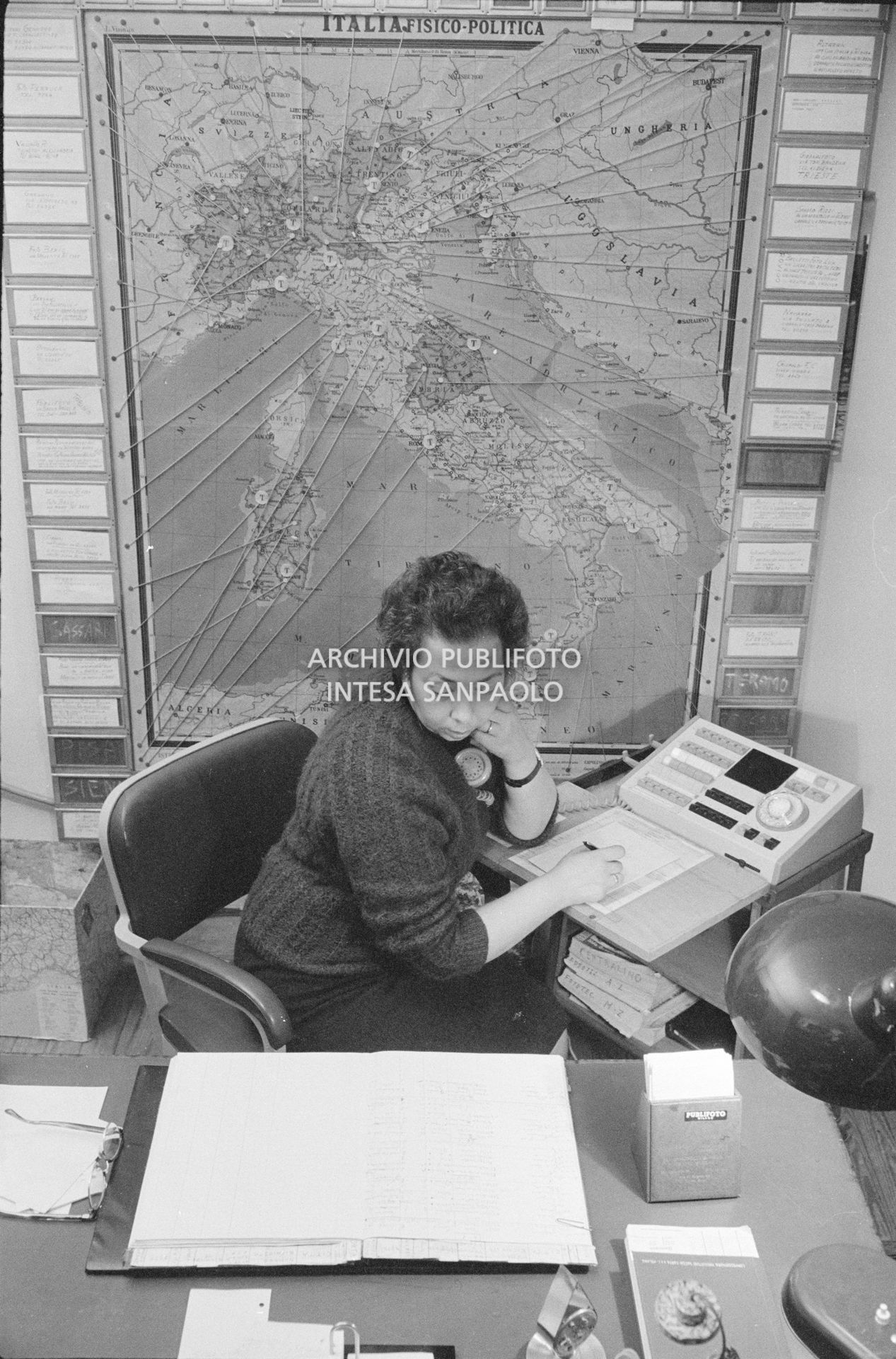 Addetta al centralino dell'agenzia Publifoto nella sede di via Bramante a Milano; dietro di lei una carta fisico politica dell'Italia con dei segnaposto numerati