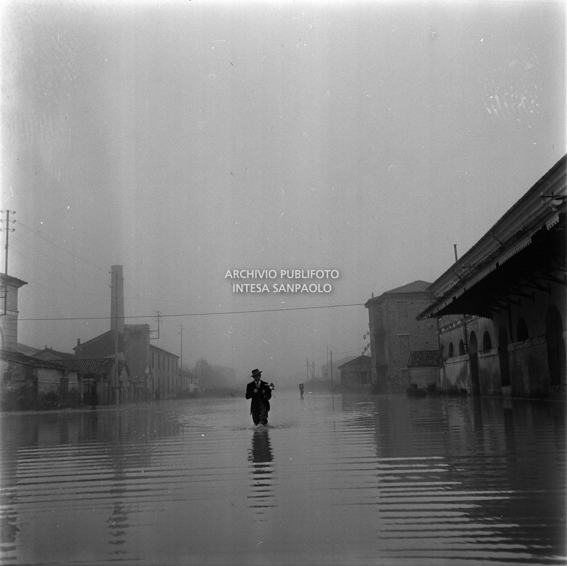 Un fotoreporter con la sua macchina fotografica cammina nell'acqua durante l'alluvione nelle campagne del Polesine; alle sue spalle in lontananza un operatore con cinepresa