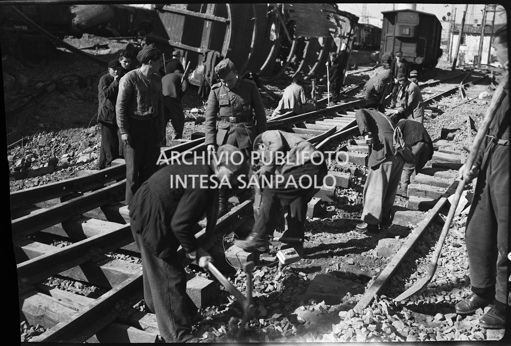 Uomini al lavoro sui binari della ferrovia, danneggiata dai bombardamenti, sotto lo sguardo di un soldato nazista che soprintende<br>26629