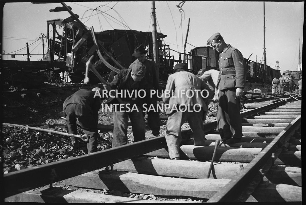 Uomini al lavoro sui binari della ferrovia, danneggiata dai bombardamenti, sotto lo sguardo di un soldato nazista che soprintende<br>26630
