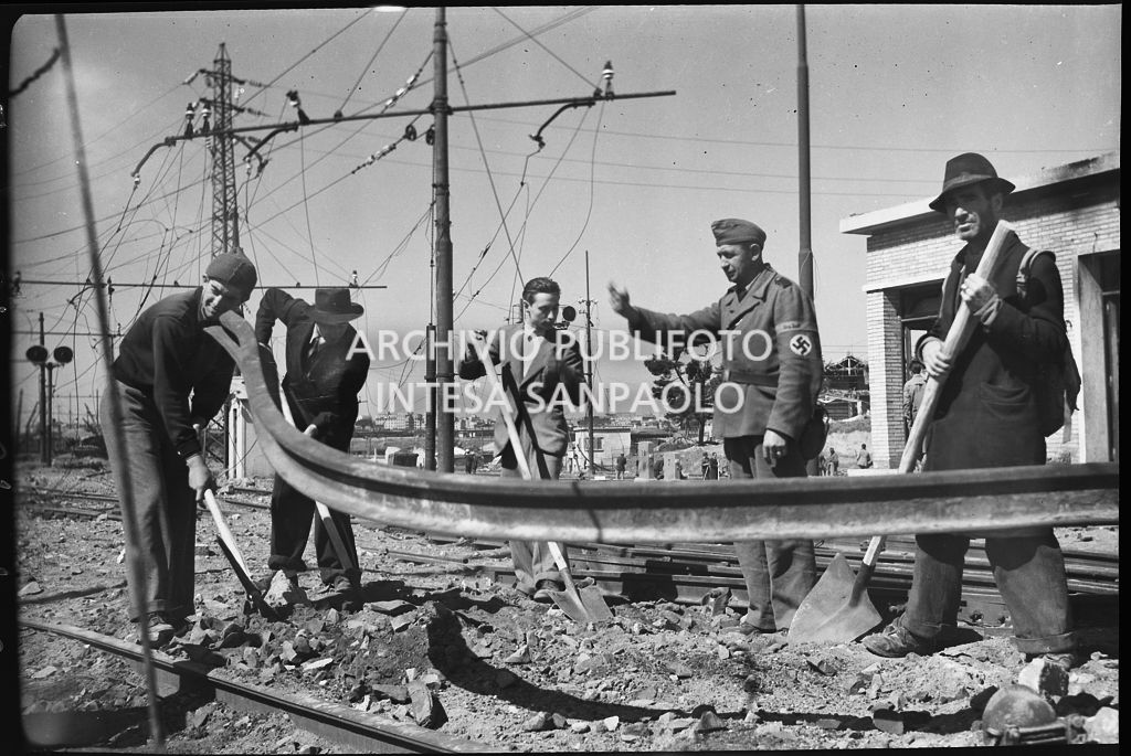 Uomini al lavoro sui binari della ferrovia, danneggiata dai bombardamenti, sotto lo sguardo di un soldato nazista che soprintende<br>26631