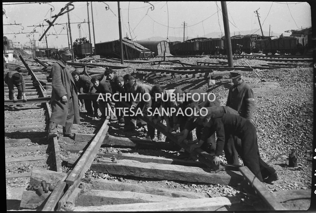 Uomini al lavoro sui binari della ferrovia, danneggiata dai bombardamenti, sotto lo sguardo di un soldato nazista che soprintende<br>26632