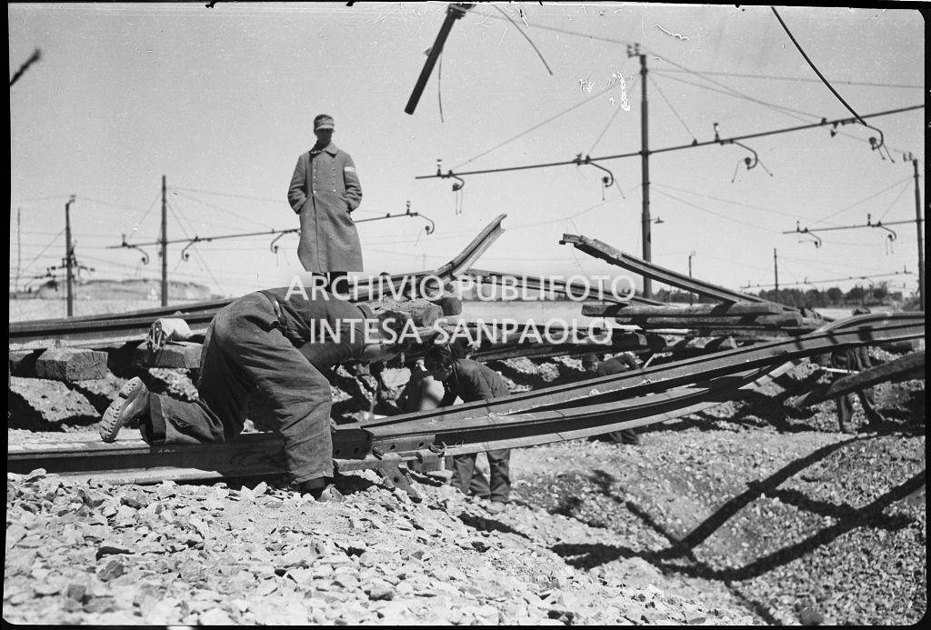 Uomini al lavoro sui binari della ferrovia, danneggiata dai bombardamenti, sotto lo sguardo di un soldato nazista che soprintende<br>26634