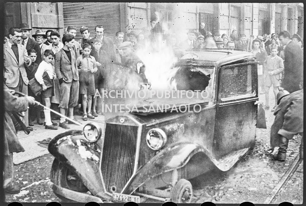 Un'automobile in fiamme; sul marciapiede persone assiepate osservano la scena<br>26638