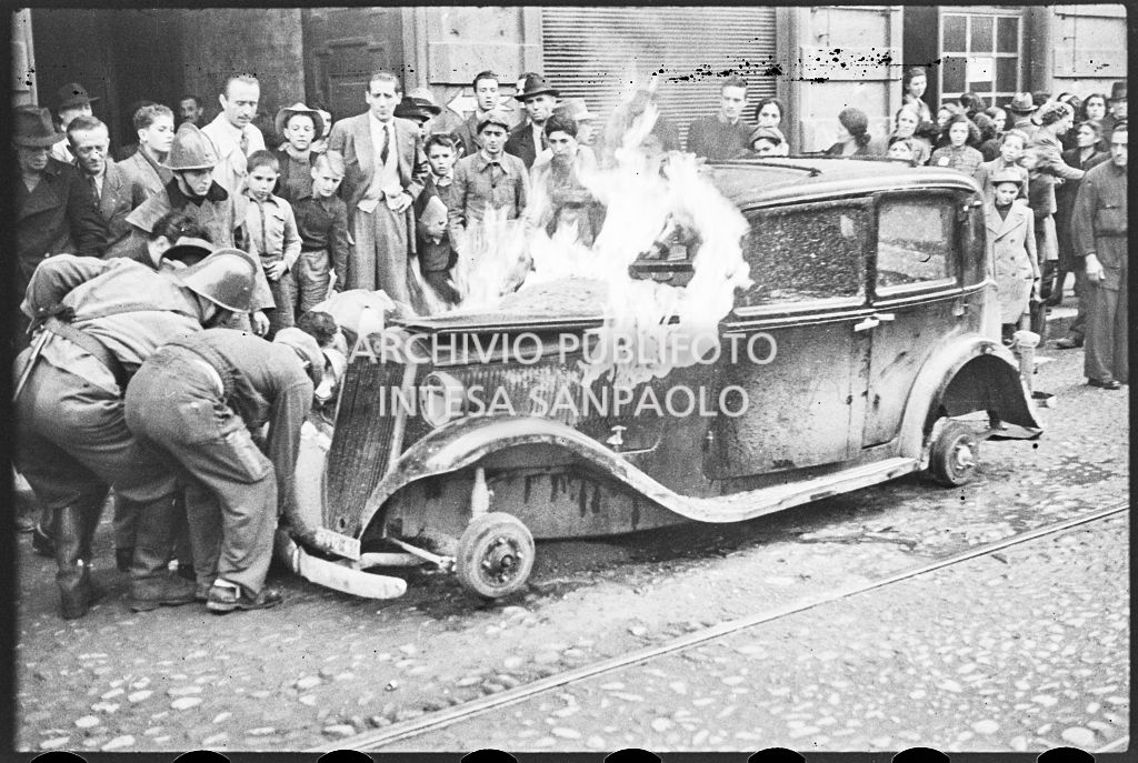 Intervento dei vigili del fuoco su un'automobile in fiamme; sul marciapiede persone assiepate osservano la scena<br>26639