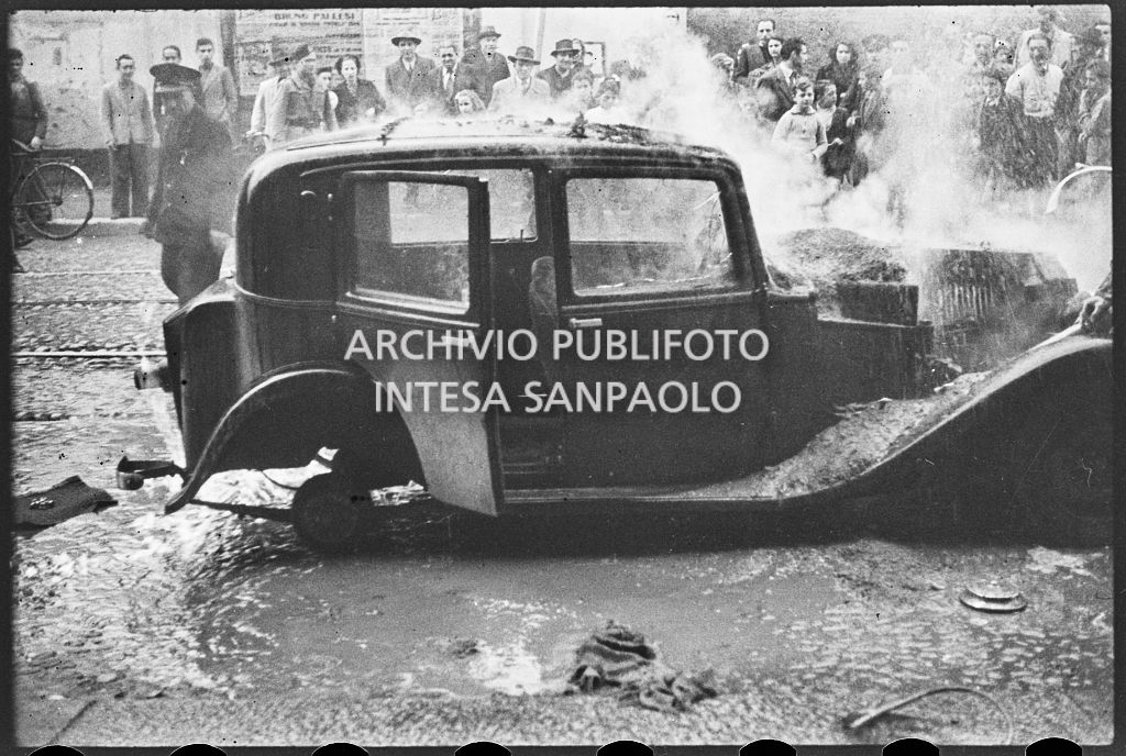 Un'automobile in fiamme; sul marciapiede persone assiepate osservano la scena<br>26640