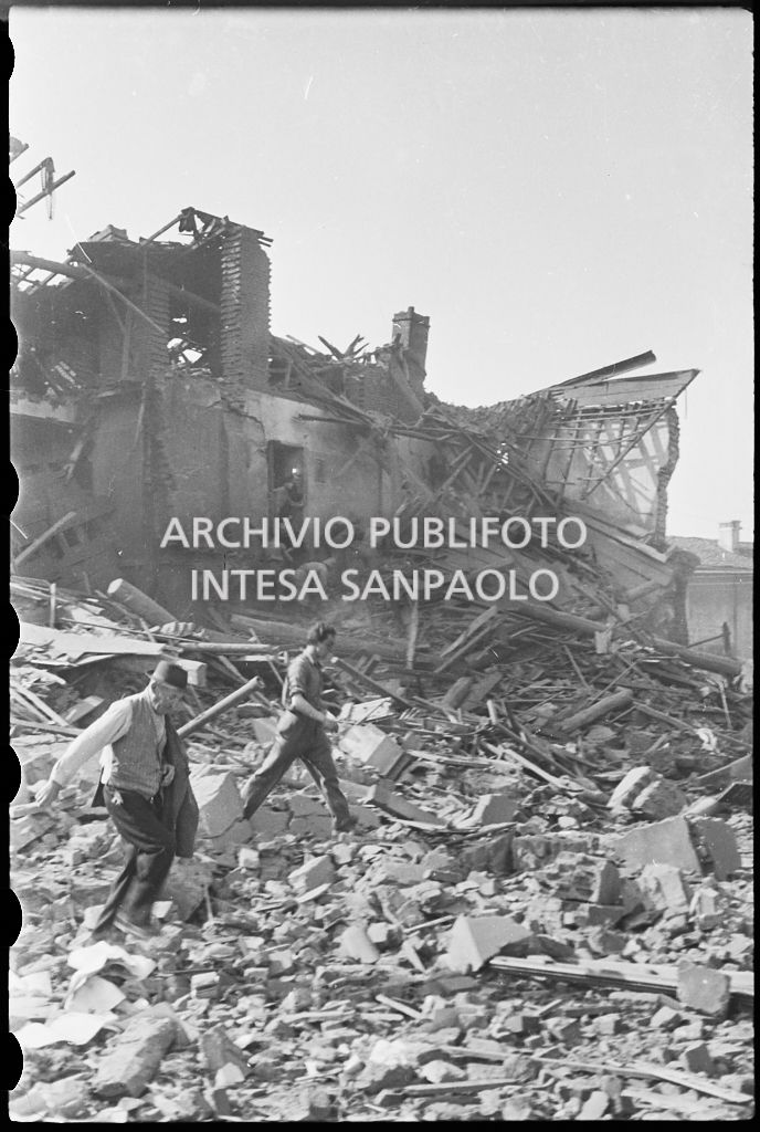 Due uomini camminano sulle macerie degli edifici bombardati; sullo sfondo intervento dei vigili del fuoco presso uno degli stabili colpiti<br>26651