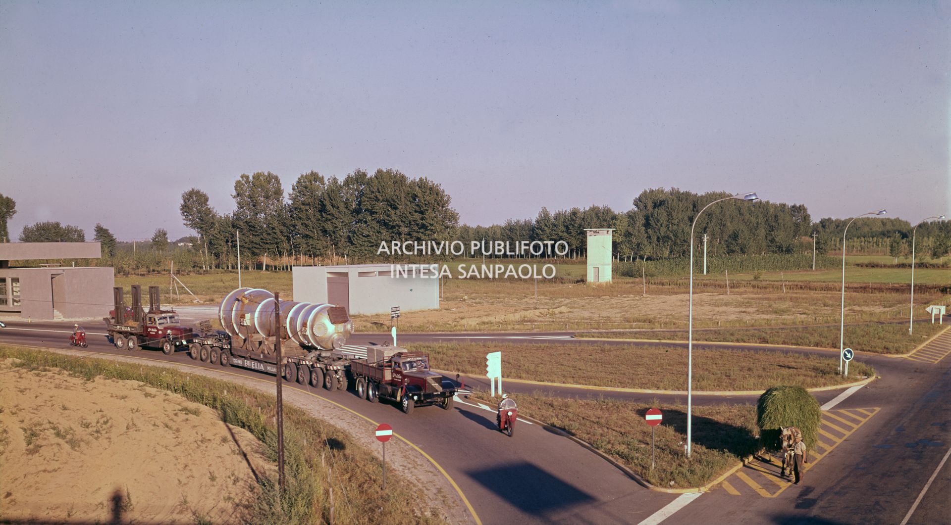 Un contadino con un carro agricolo nei pressi di un uscita autostradale, visibile un convoglio speciale fermo all'uscita