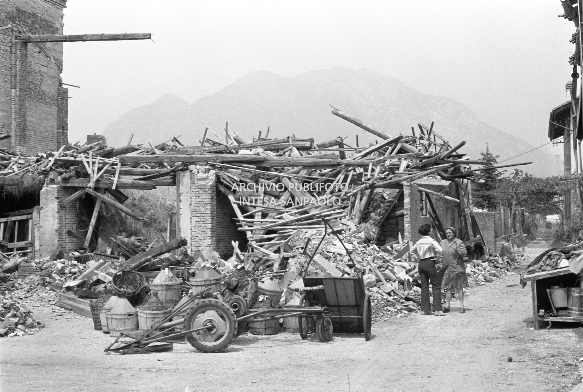 Due donne in località colpita dal terremoto, visibili le macerie di alcune costruzioni e alcune damigiane di vino
